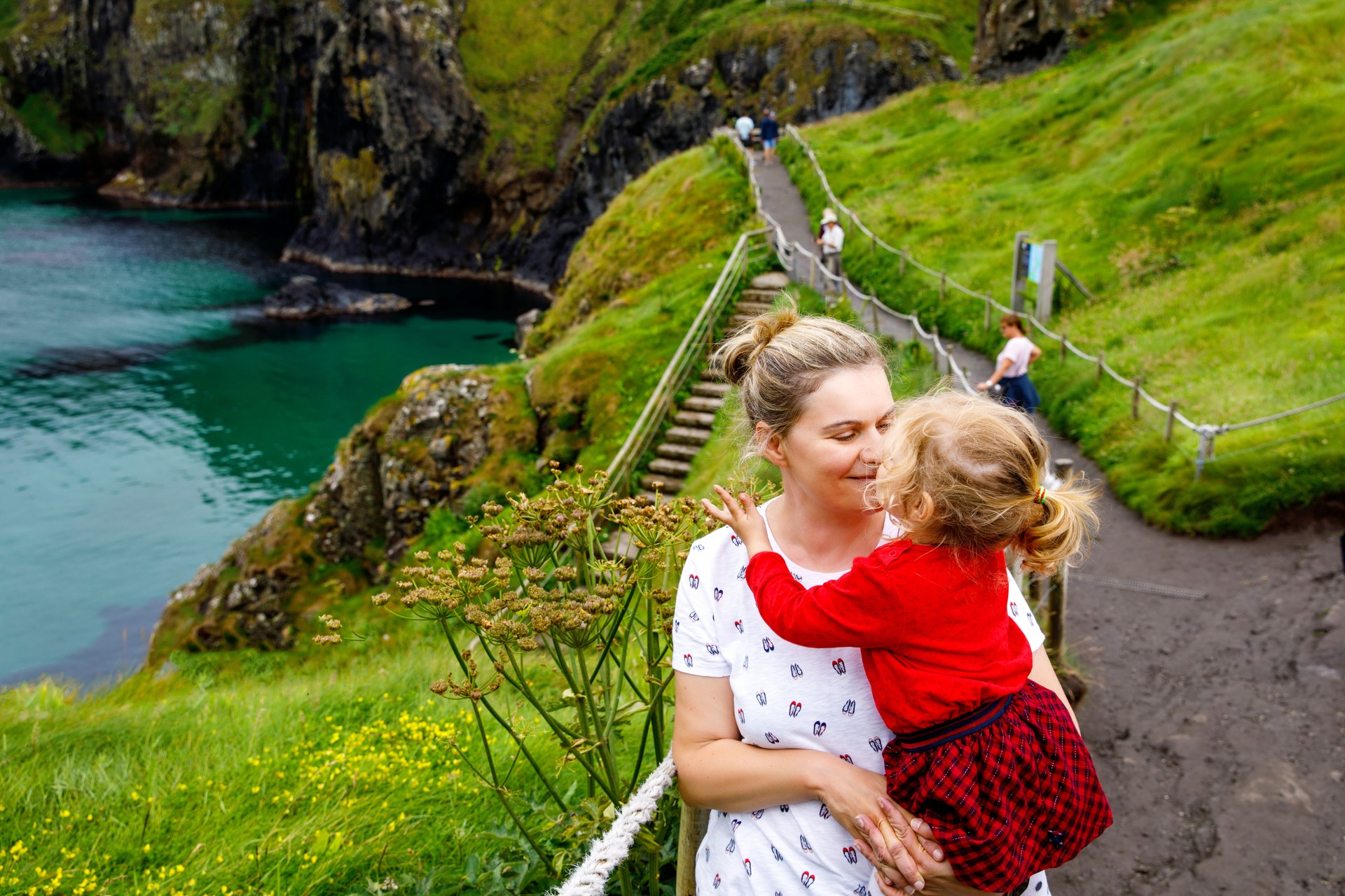 Toddler girl and mother on Carrick-a-Rede Rope Bridge, famous rope bridge near Ballintoy, Northern Ireland on Irish coastline. Family of child and woman on bridge to small island on cloudy day.